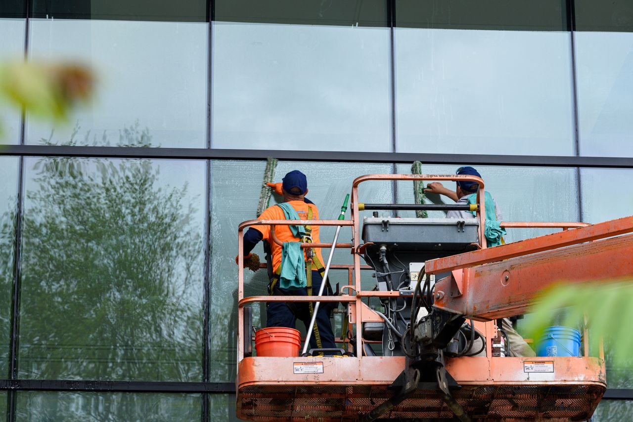 Two workers washing windows on a glass building facade from a lift platform, improving the building’s appearance.